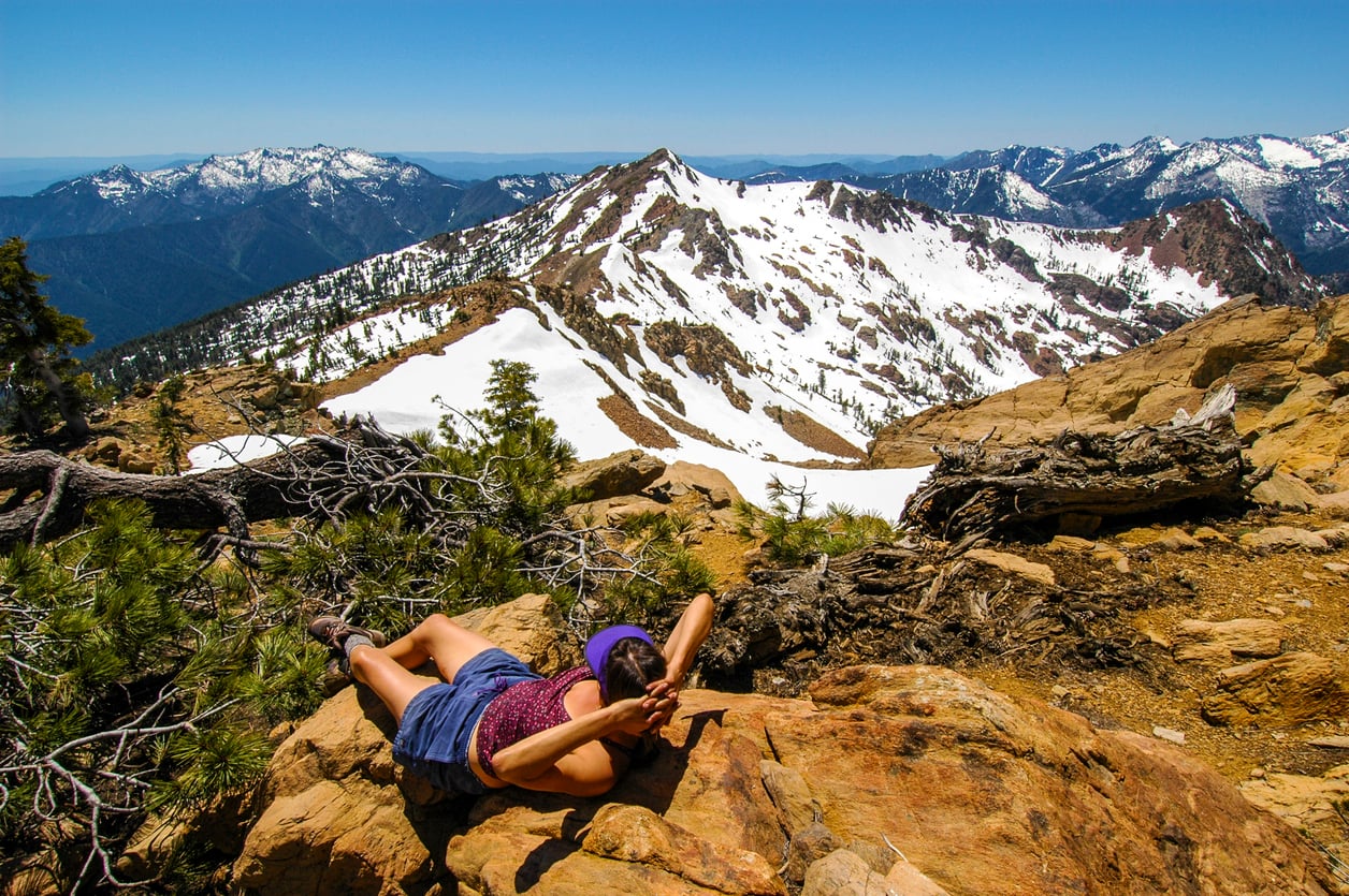 A person in a purple outfit lies on a rocky outcrop, relaxing with hands behind their head, overlooking snow-capped mountains under a clear blue sky.