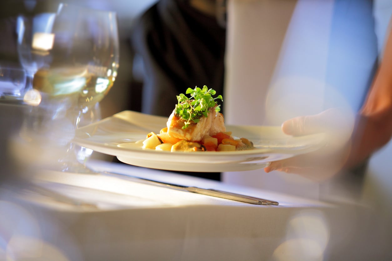 A person serves a white plate with a gourmet dish, including salmon, vegetables, and microgreens, in a fine dining setting with soft lighting and blurred glassware in the foreground.