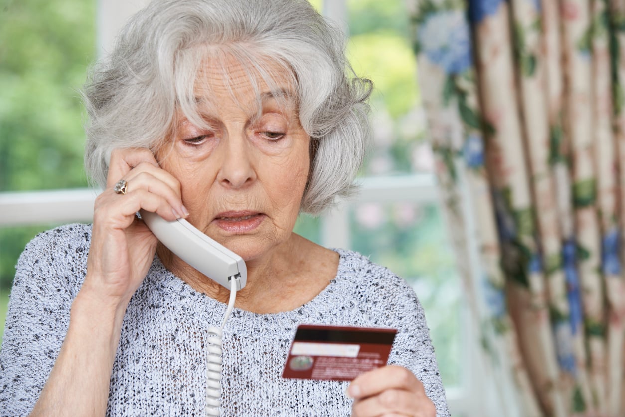 An elderly woman with gray hair holds a phone to her ear and looks worriedly at a credit card in her hand, sitting indoors near a window with floral curtains.