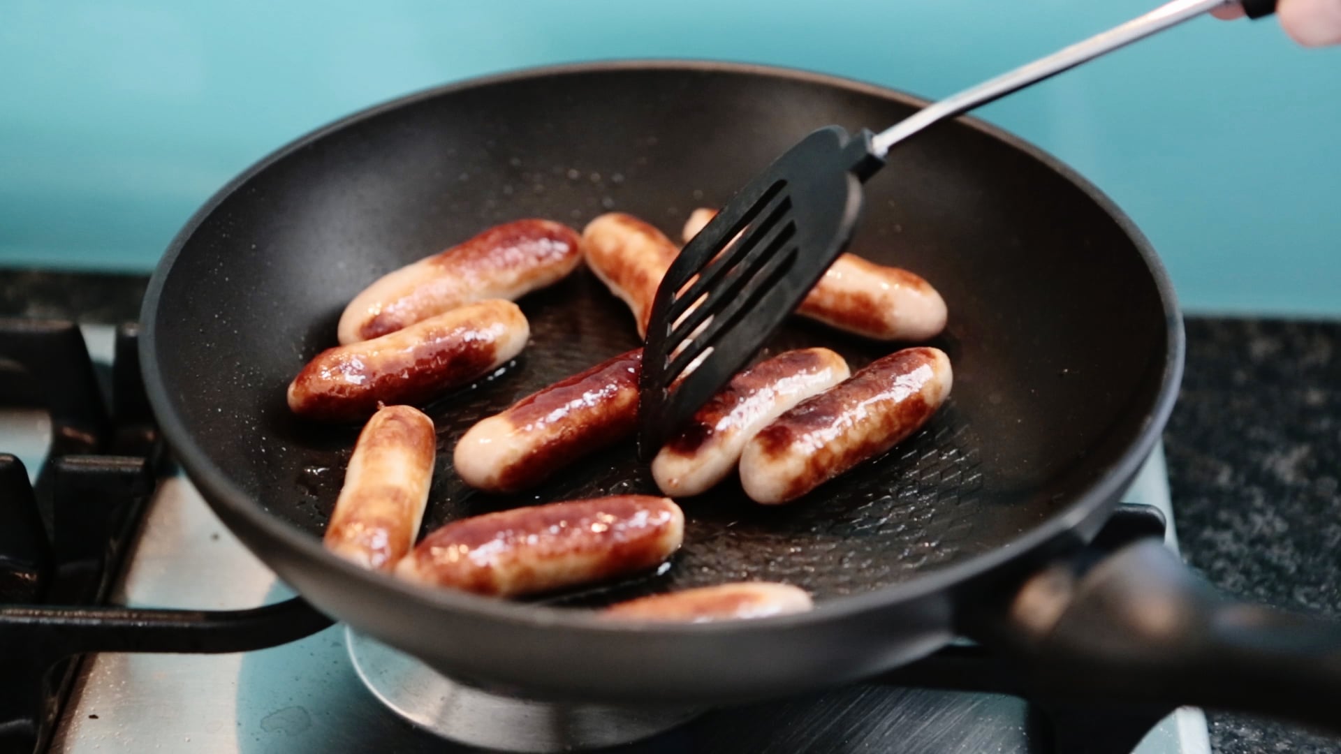 A person uses a black spatula to cook several browned sausages in a nonstick frying pan on a stovetop with a blue background.