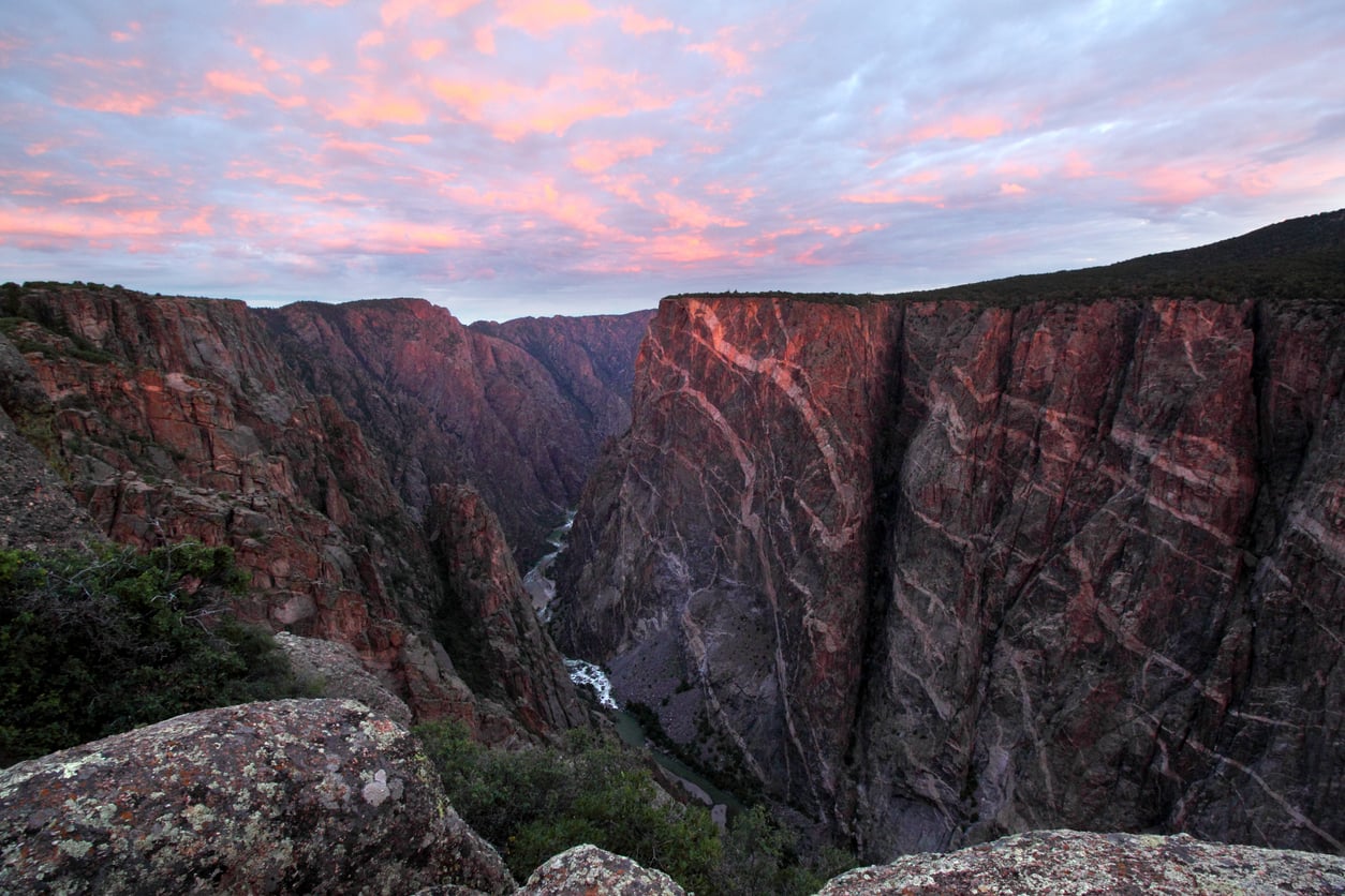 View of a deep, steep canyon with rugged, multicolored rock walls under a sunset sky with pink clouds. A narrow river flows at the canyon’s base, surrounded by dramatic cliffs and sparse vegetation.