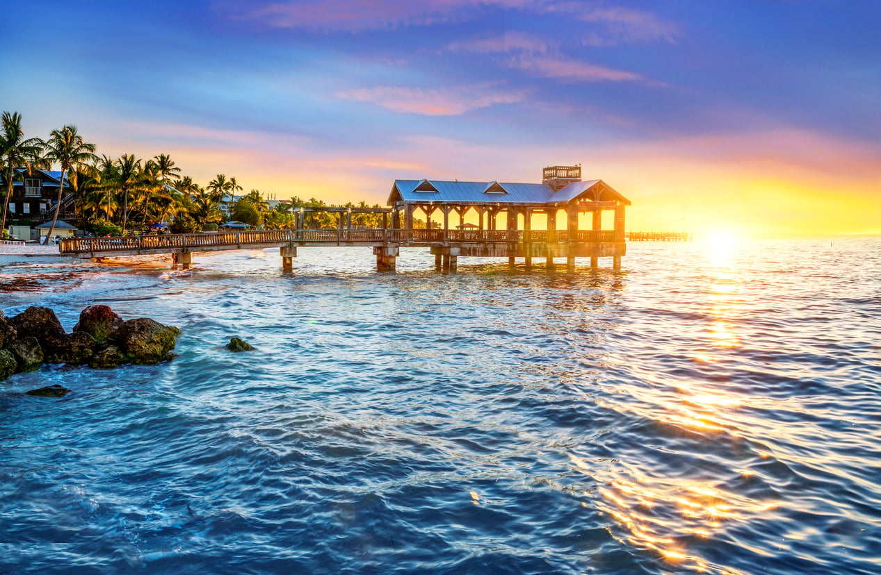 A vibrant sunset over a wooden pier stretching into the ocean, with waves reflecting colorful skies and palm trees lining the shore.