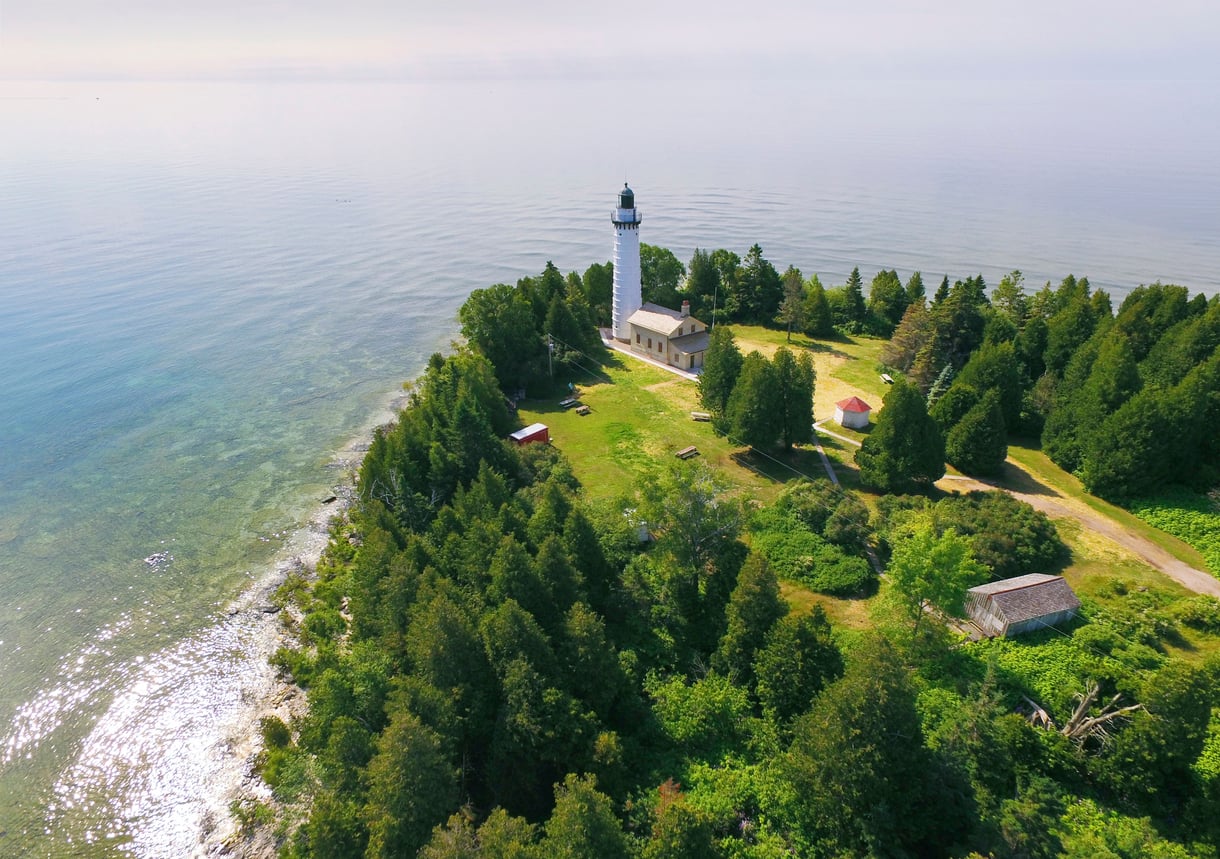 Aerial view of a lighthouse near the shore, surrounded by trees and a few small buildings, overlooking calm, clear water.