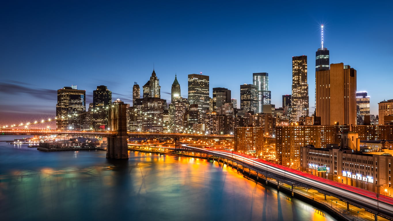 New York City skyline at dusk with illuminated skyscrapers, Brooklyn Bridge, and light trails from cars reflecting on the water.