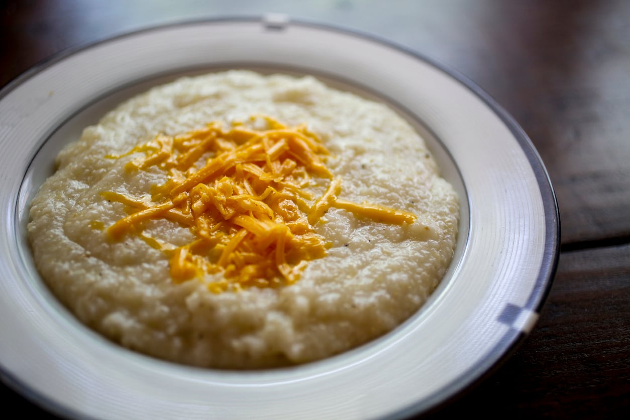 A white plate with creamy grits topped with shredded cheddar cheese, sitting on a dark wooden table.