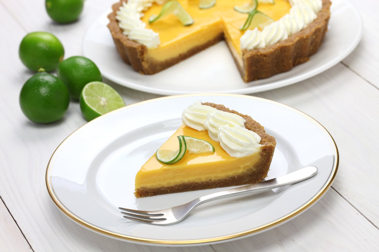 A slice of key lime pie topped with whipped cream and lime slices sits on a white plate with a fork. The whole pie and fresh limes are in the background on a white wooden table.