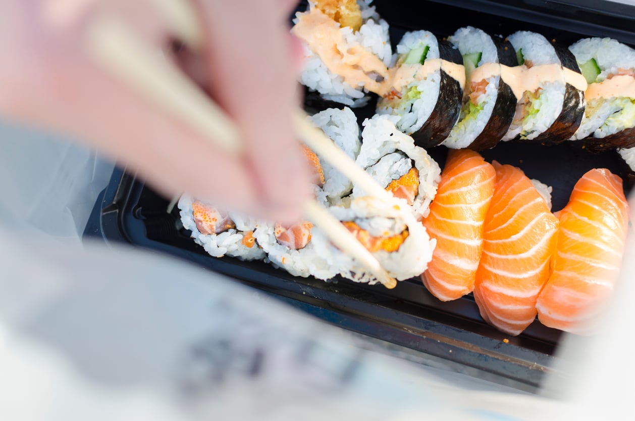 A hand uses chopsticks to pick up a sushi roll from a black tray containing various sushi, including salmon nigiri and different types of rolls—perfect examples of popular foods to order at any Japanese restaurant.