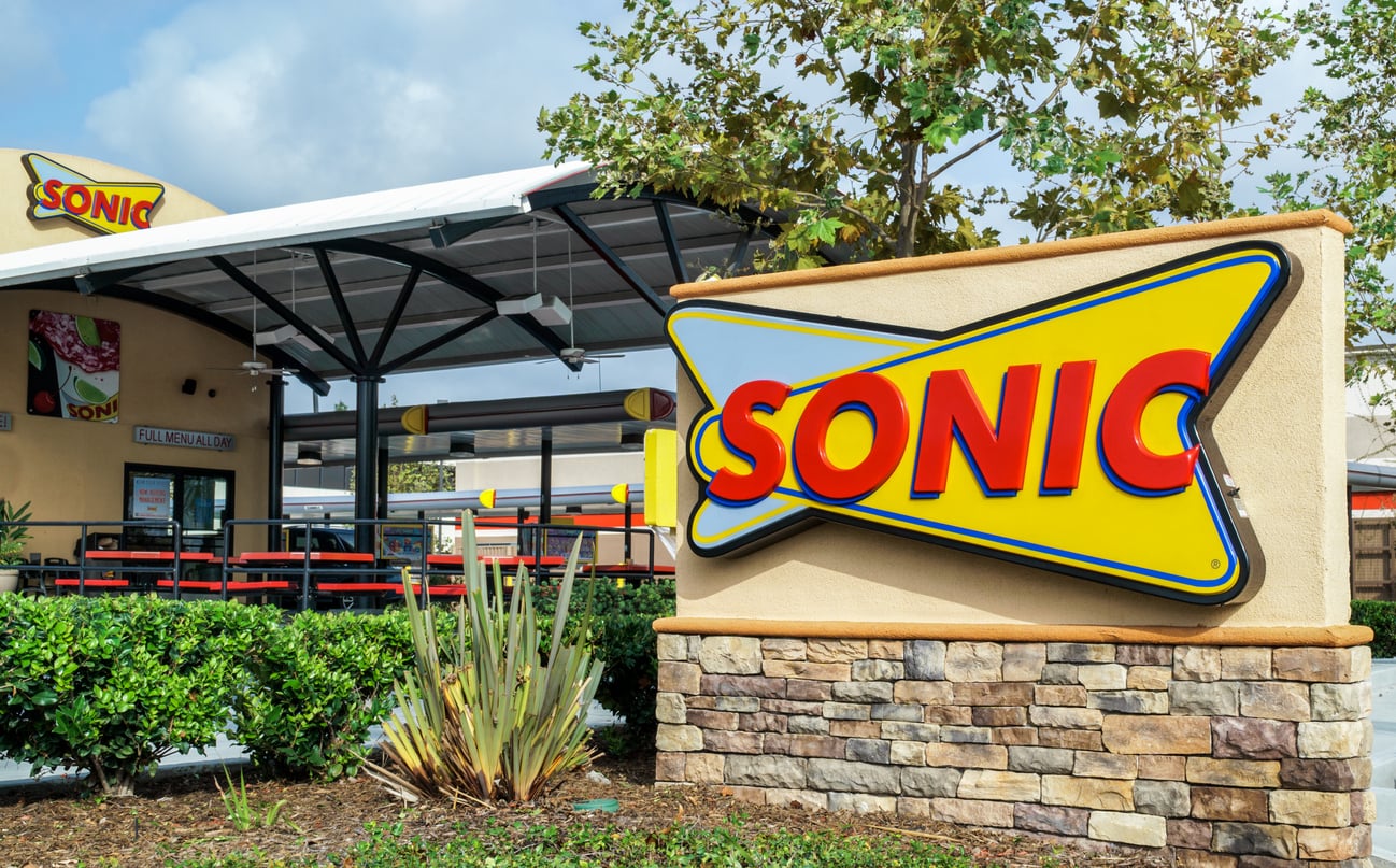 A Sonic Drive-In restaurant exterior with a large yellow SONIC sign in front, outdoor seating, drive-in stalls, greenery, and a stone planter.