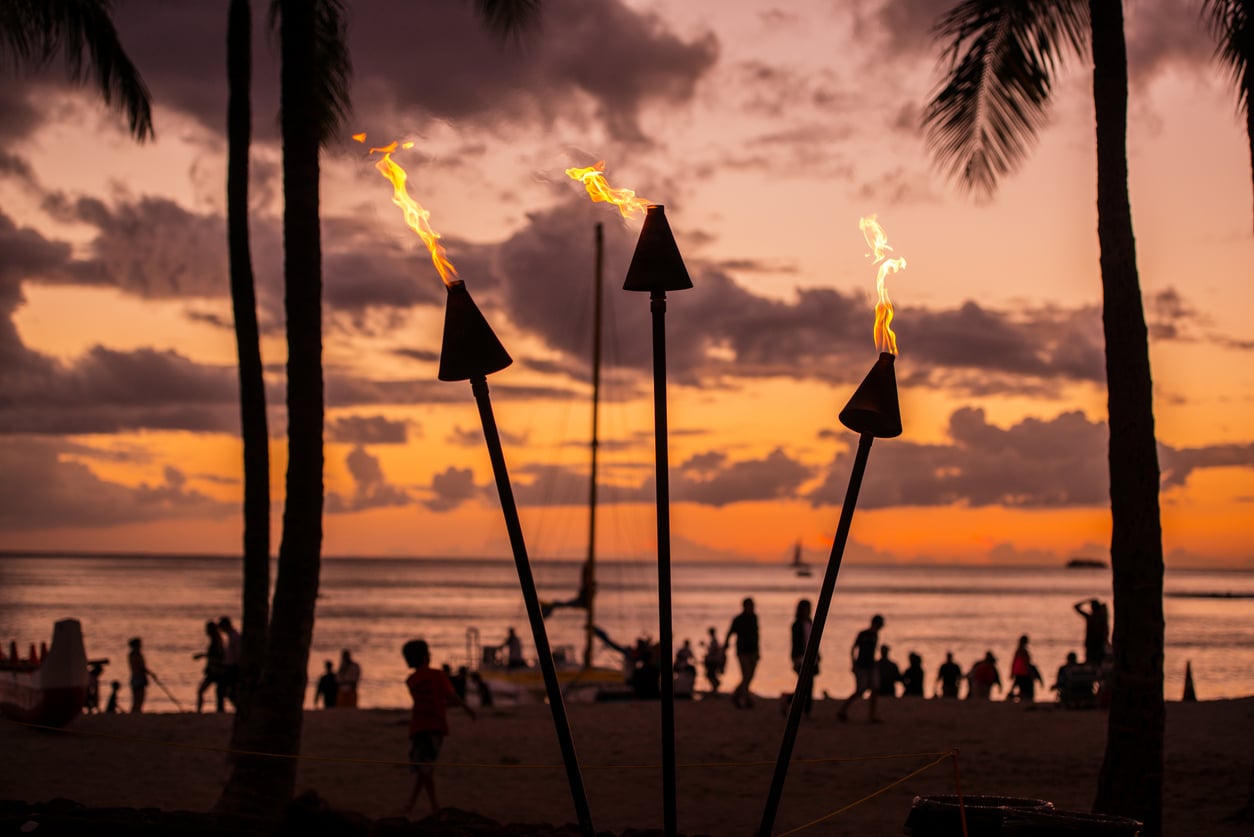 Three tiki torches with small flames stand on a beach at sunset, evoking the relaxed spirit of vacation; silhouettes of people, palm trees, and a sailboat are visible against an orange and purple sky.