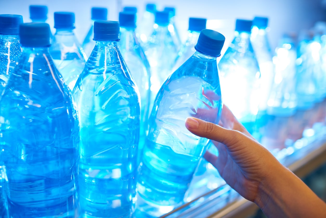 A person’s hand is grabbing a plastic bottle of water from a brightly lit refrigerator shelf filled with other bottles of water, all with blue caps.