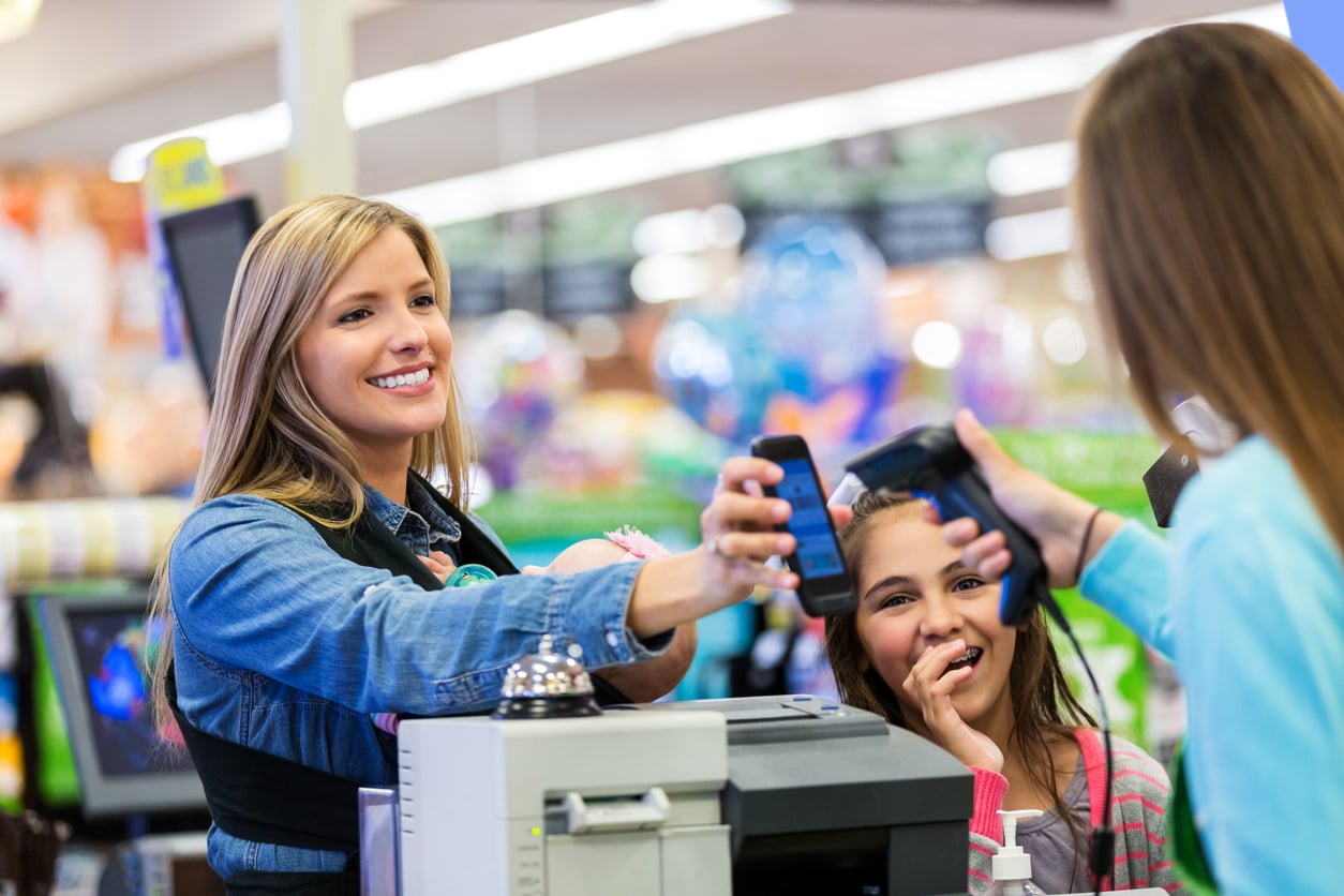 A smiling cashier scans a customer’s smartphone at a checkout counter while a young girl stands nearby, smiling, in a brightly lit store.