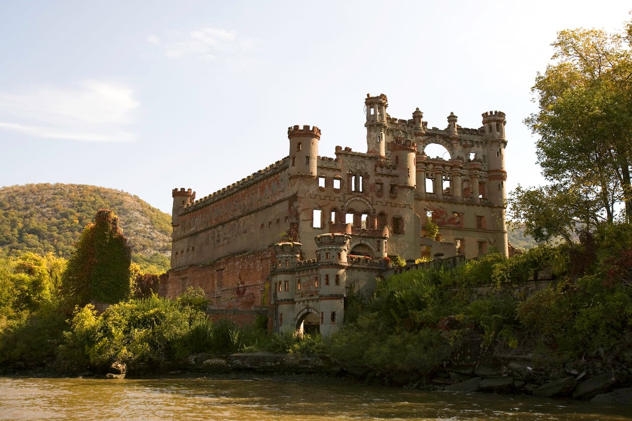 Ruins of a large, castle-like brick building stand by the water, surrounded by trees and greenery, with a hill visible in the background under a partly cloudy sky—a silent testament to the era of grand castles.