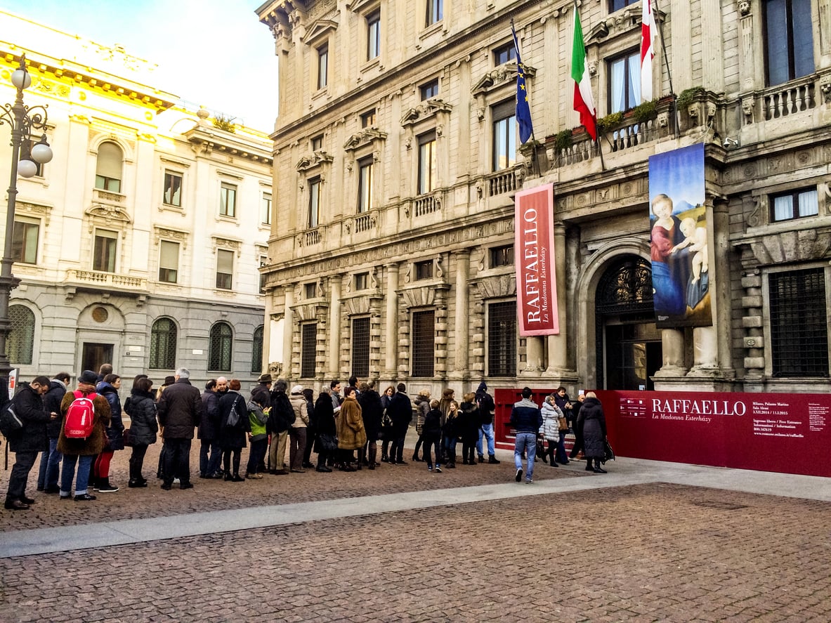 A long line of people waits outside a grand stone building with “Raffaello” exhibition banners and flags hanging above the entrance on a sunny day—a reminder that not booking tickets in advance can be one of the common vacation mistakes.