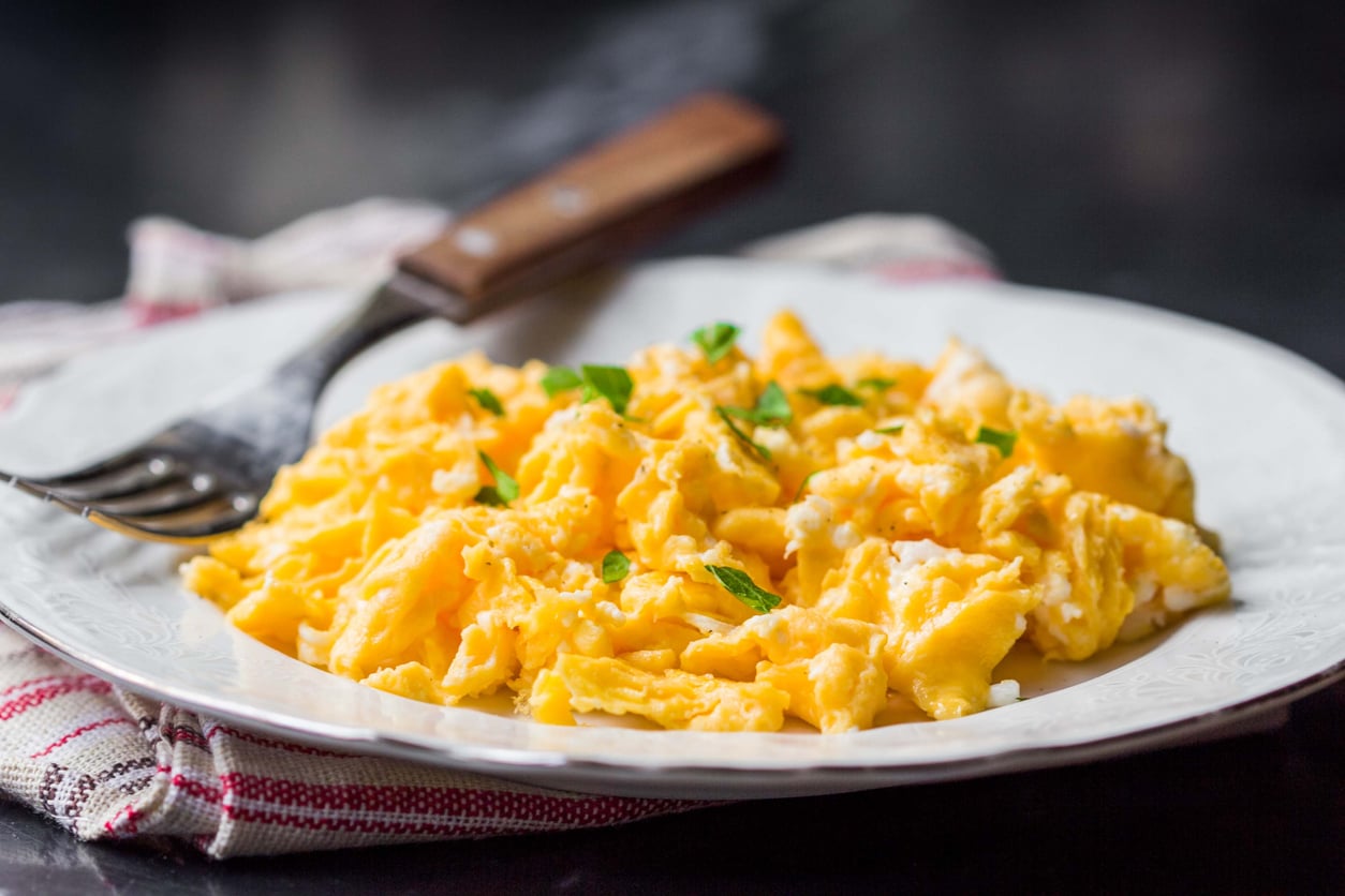 Scrambled eggs, one of the most beloved breakfast foods, are garnished with chopped herbs on a white plate, next to a fork with a brown handle on a red and white cloth.