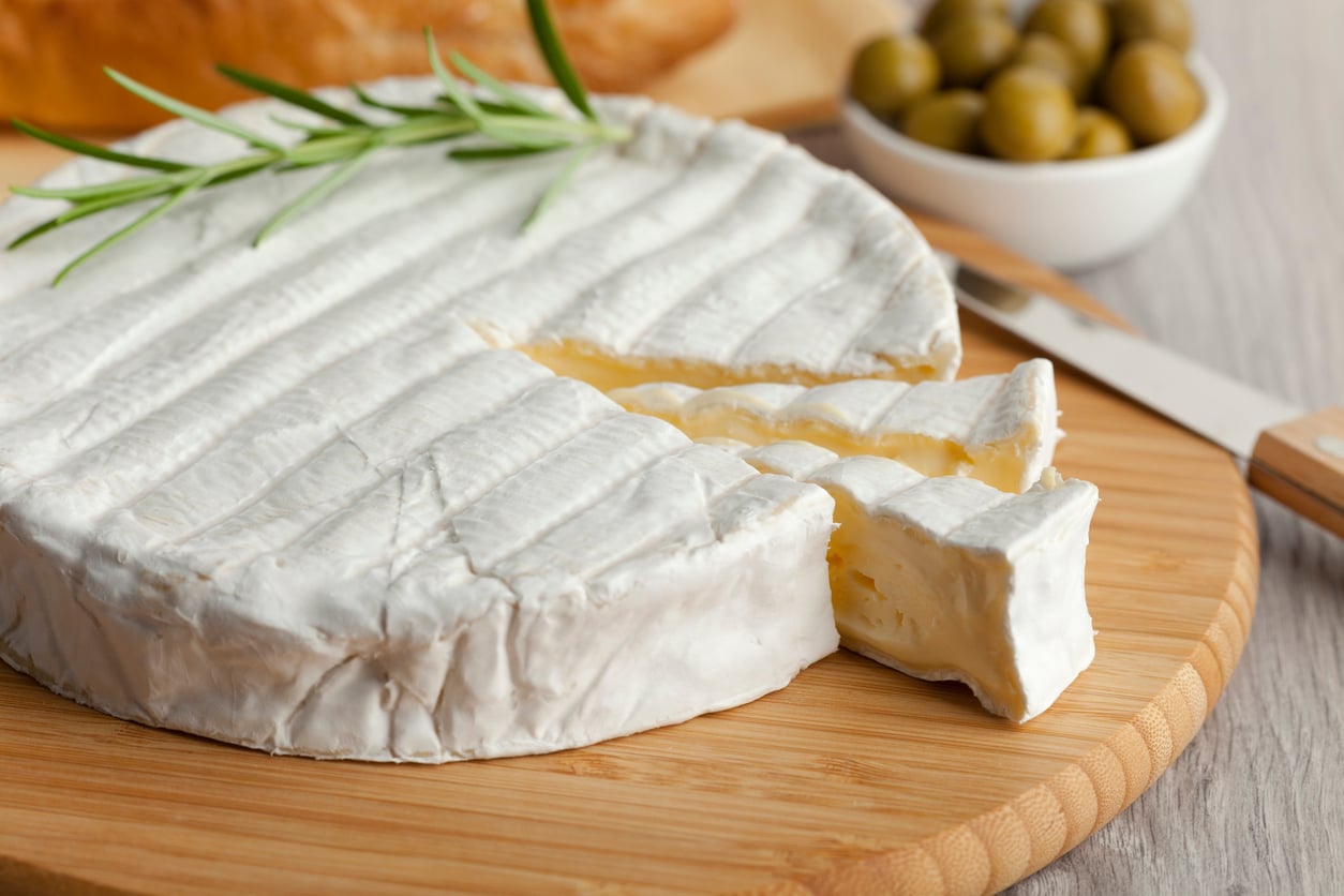 A round wheel of soft brie cheese with a wedge cut out sits on a wooden board, garnished with rosemary—a delicious example of everyday foods that used to be considered fancy. A knife, olives, and bread are in the background.
