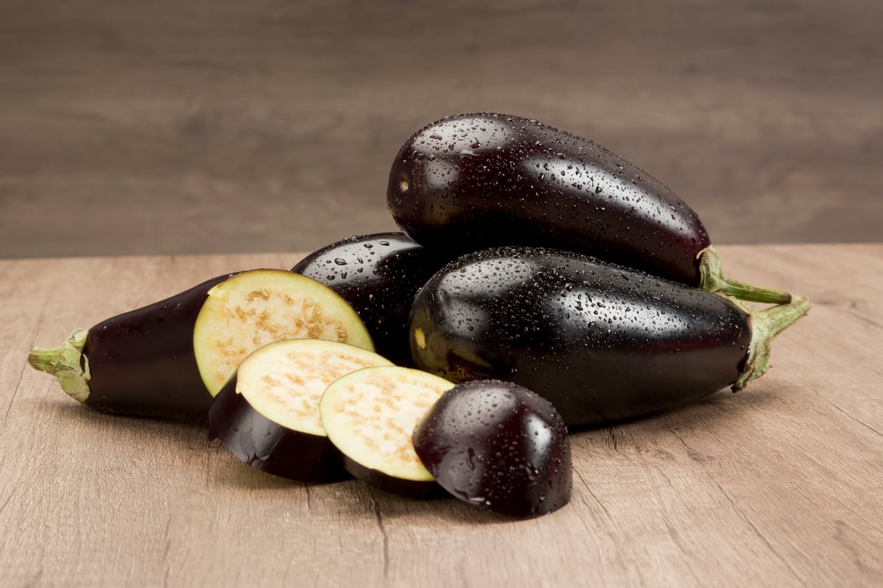 Three whole eggplants rest on a wooden surface, with two round slices and one half piece in front. The eggplants, often considered among the foods people learn to appreciate later in life, are dark purple with drops of water on their skin. The background is brown and out of focus.