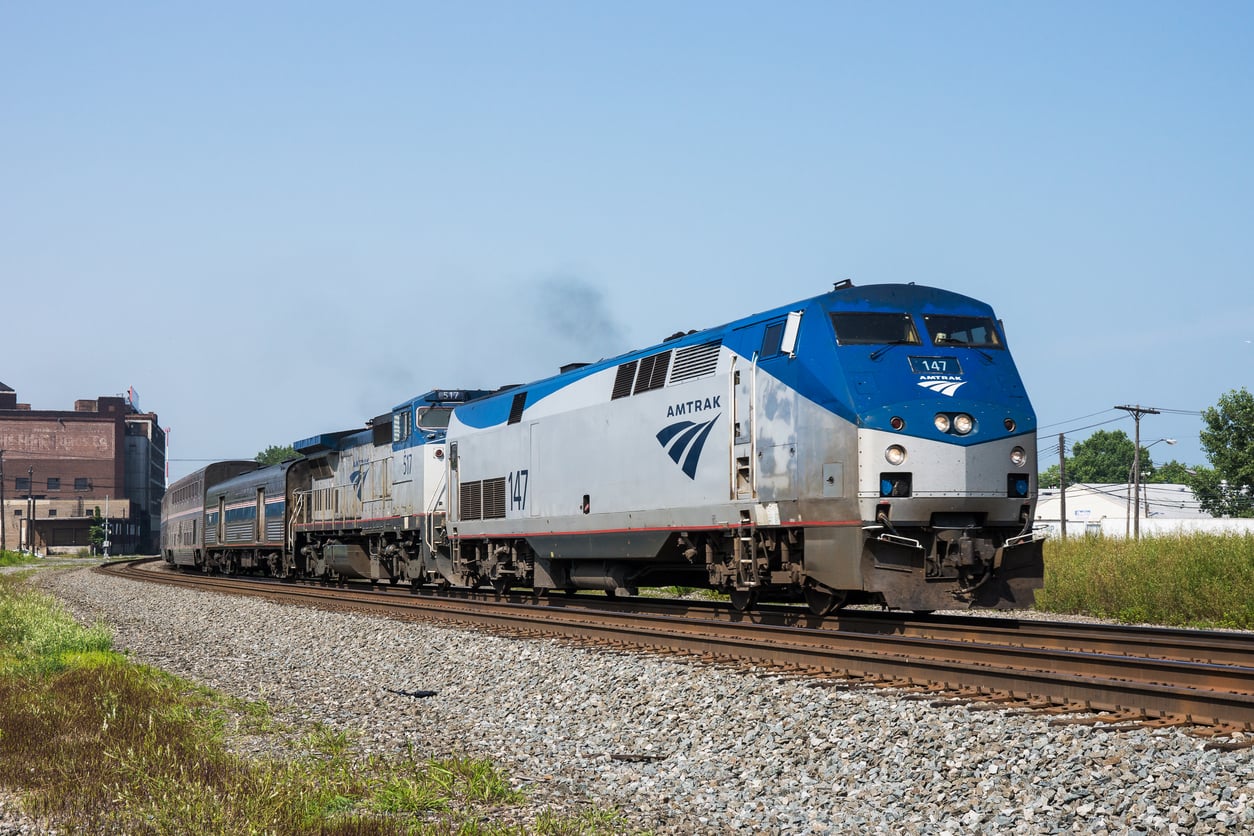 An Amtrak passenger train travels down a curved railroad track on a clear day, with buildings and green trees visible in the background.