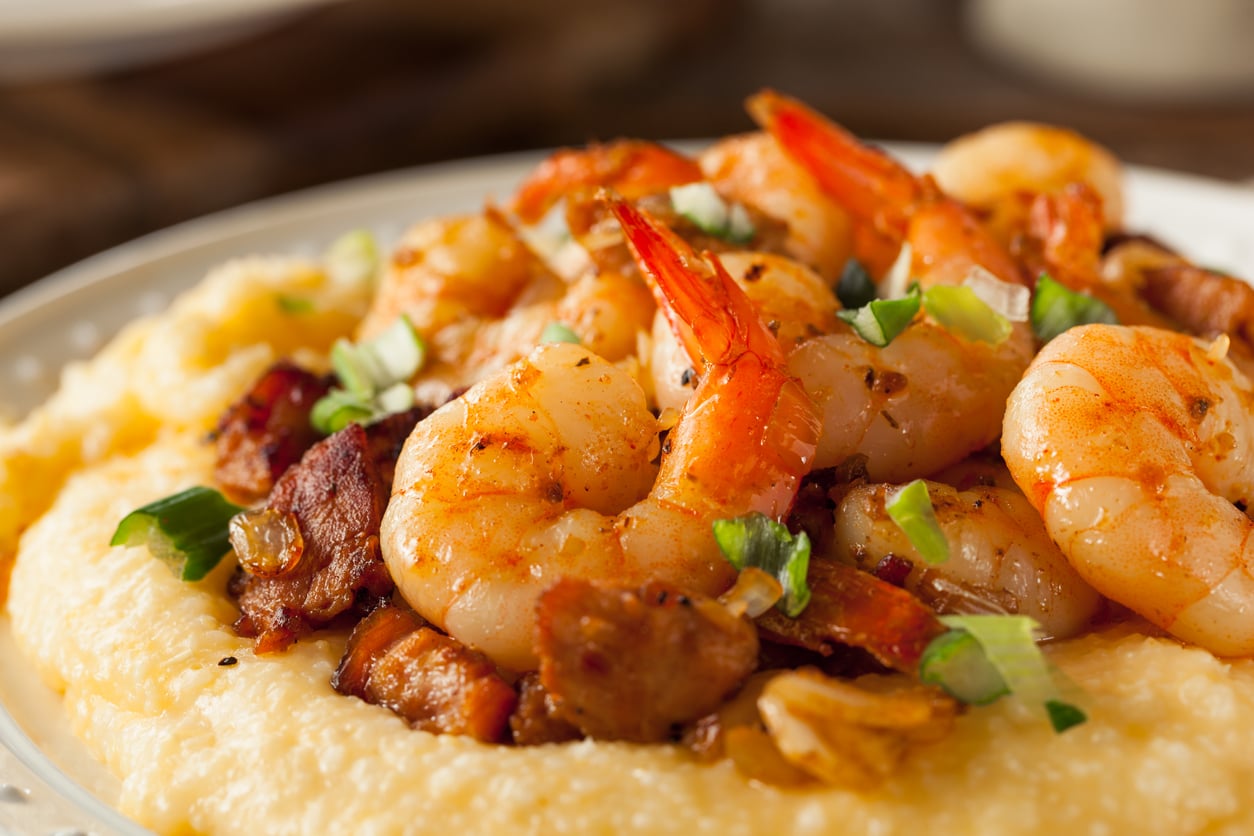 A close-up of shrimp and pieces of bacon served over creamy yellow grits, garnished with chopped green onions on a white plate.
