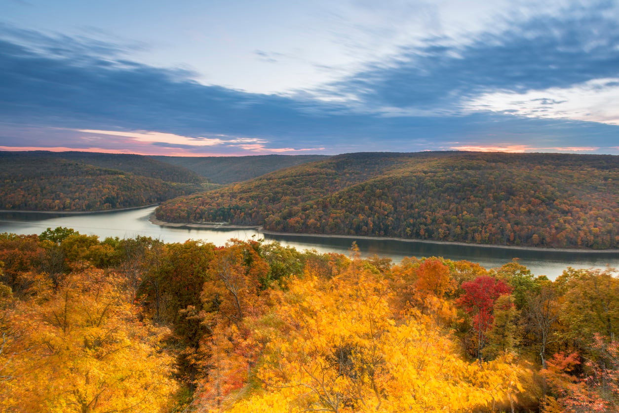 A wide river curves between hills covered in autumn foliage, with vibrant yellow, orange, and red trees in the foreground and a cloudy sky at sunset—making this breathtaking landscape one of the region’s most idyllic hiking destinations.