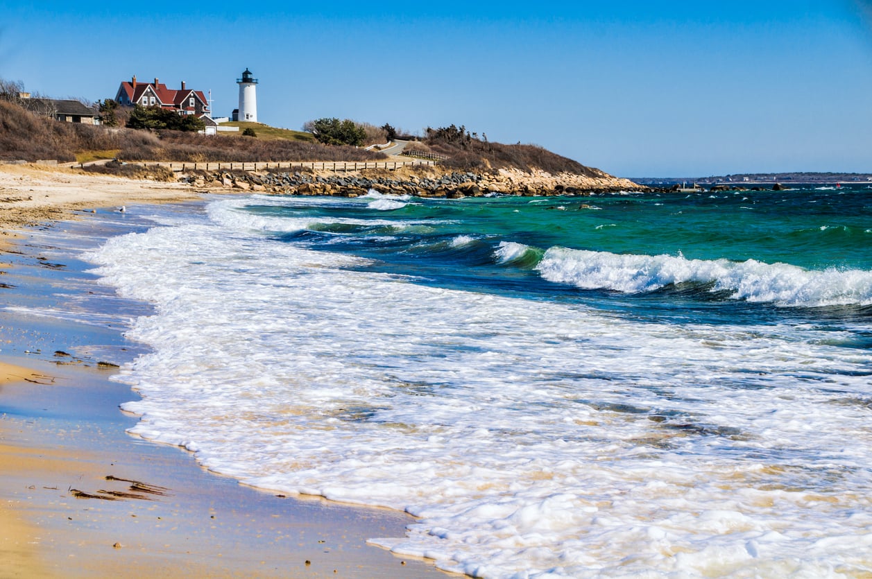 Waves crash onto a sandy beach under a clear blue sky. A white lighthouse and house sit on a grassy hill in the background, near a wooden fence.