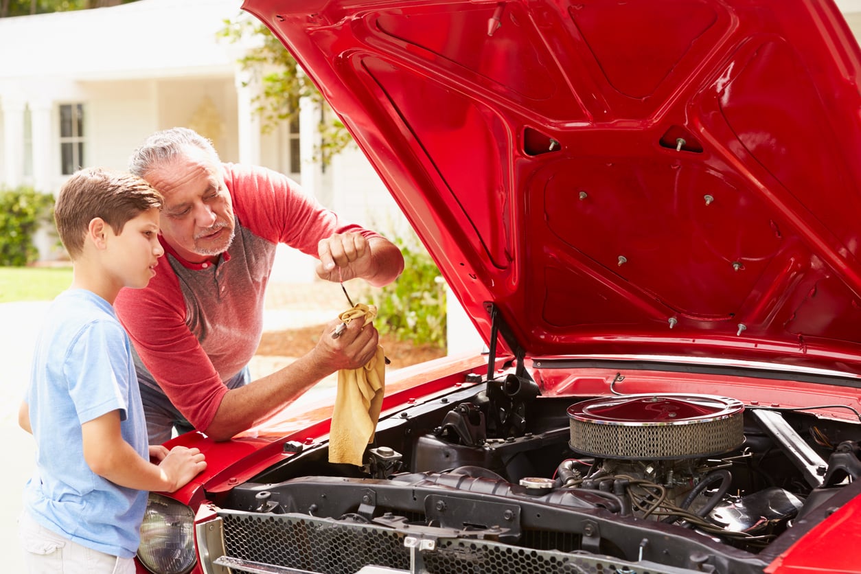 An older man shows a young boy how to check a car engine’s oil with a dipstick. They stand by a classic red car with the hood open, outside a house.
