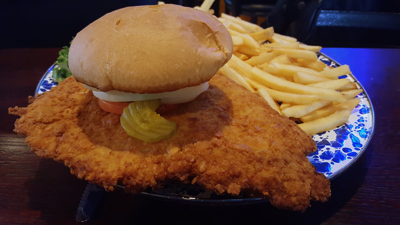 A large breaded fried pork tenderloin sandwich with pickles, tomato, and onion, served on a bun with a side of thin French fries on a blue and white plate.