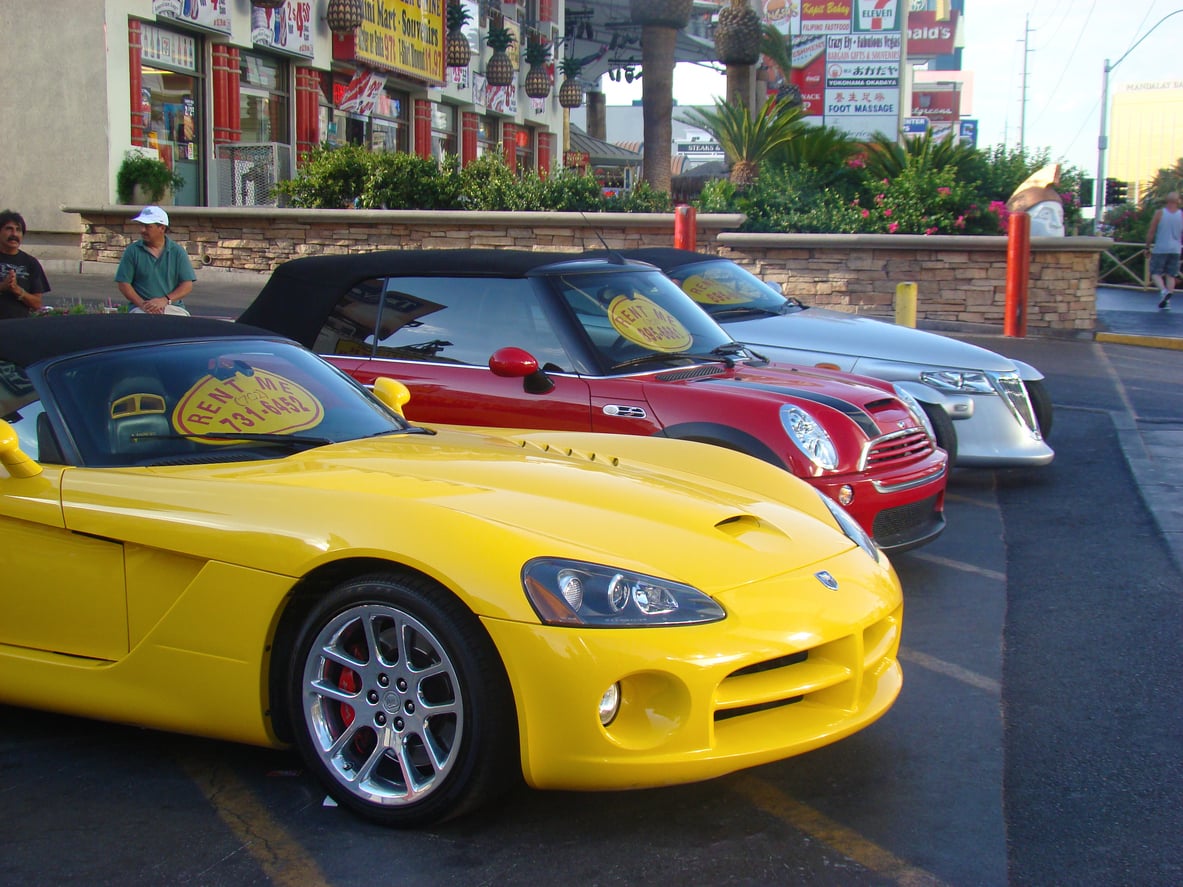 Three cars—a yellow sports car, a red convertible, and a silver sedan—are parked side by side outside a shop with signs. A “Rent Me” sign is visible on the yellow and red cars’ windshields.