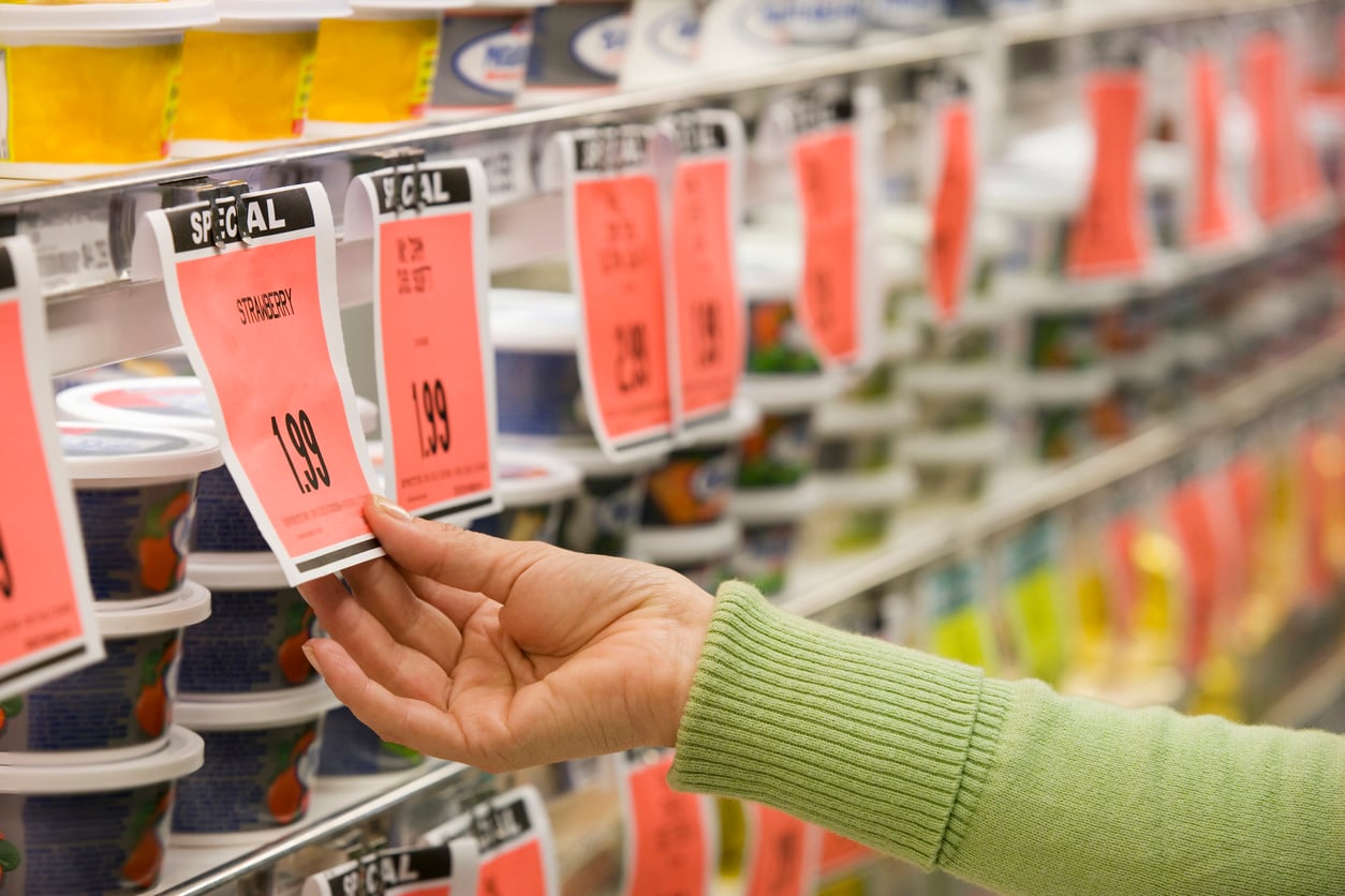 A person in a green sweater, demonstrating mindful grocery habits, reaches for a yogurt container on a grocery store shelf, where multiple items are marked with large orange sale tags.