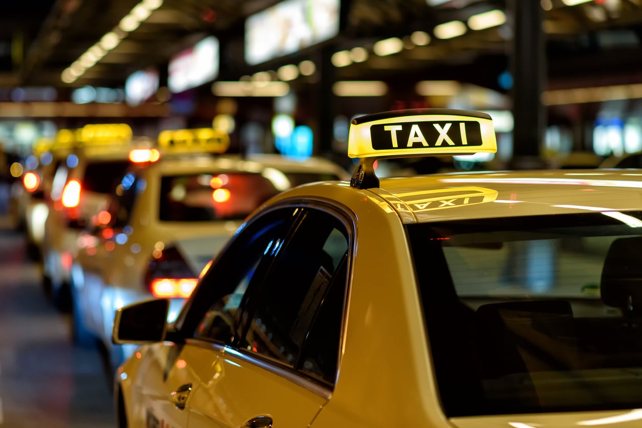 A row of taxis with illuminated “TAXI” signs waits in a line at night, likely at a busy transportation hub or airport, with blurred lights and other vehicles in the background.