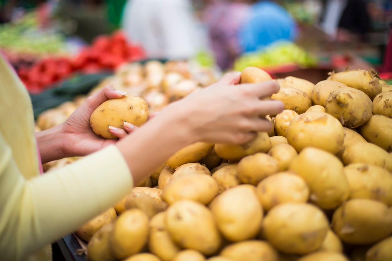 Person selecting fresh potatoes from a large pile at an outdoor market, with blurred fruits and vegetables visible in the background.