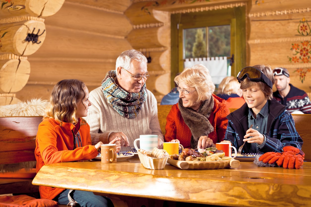 Four people, two older adults and two children, sit at a wooden table in a cozy log cabin, enjoying food and drinks together. The atmosphere is warm and cheerful, with winter clothes and decorations visible.
