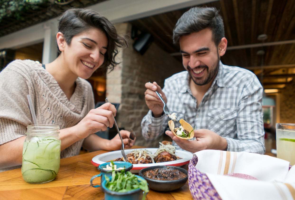 Two people sitting at a table, smiling and eating tacos together. There are plates of food and drinks on the wooden table, and the setting appears to be a cozy restaurant with warm lighting.