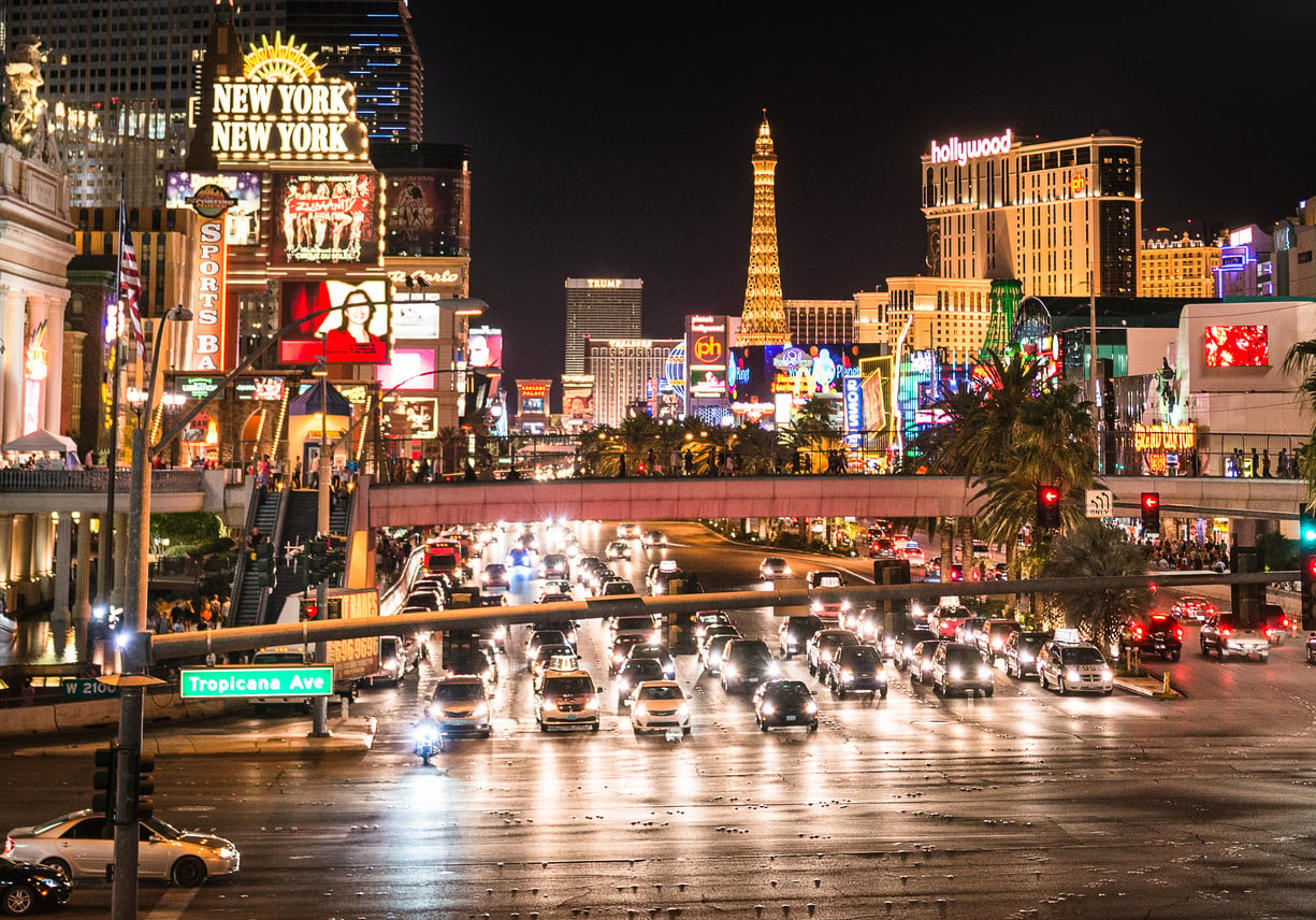 Las Vegas Strip at night with bright lights, neon signs, heavy traffic, palm trees, and replicas of famous landmarks like the Eiffel Tower and New York skyscrapers.