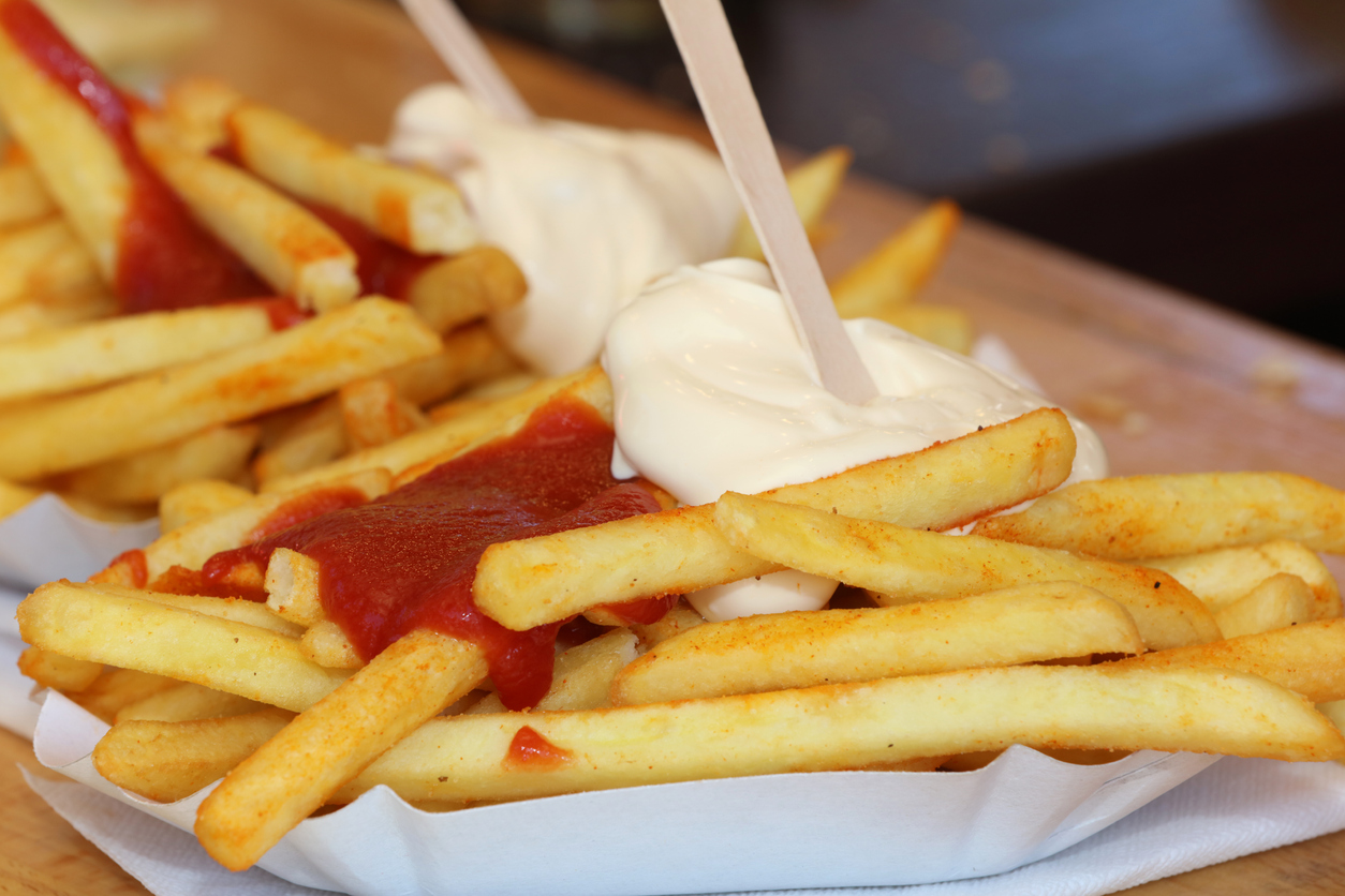 French fries, one of the most beloved comfort foods, are served in a paper tray, topped with ketchup and mayonnaise, with wooden forks inserted for eating.