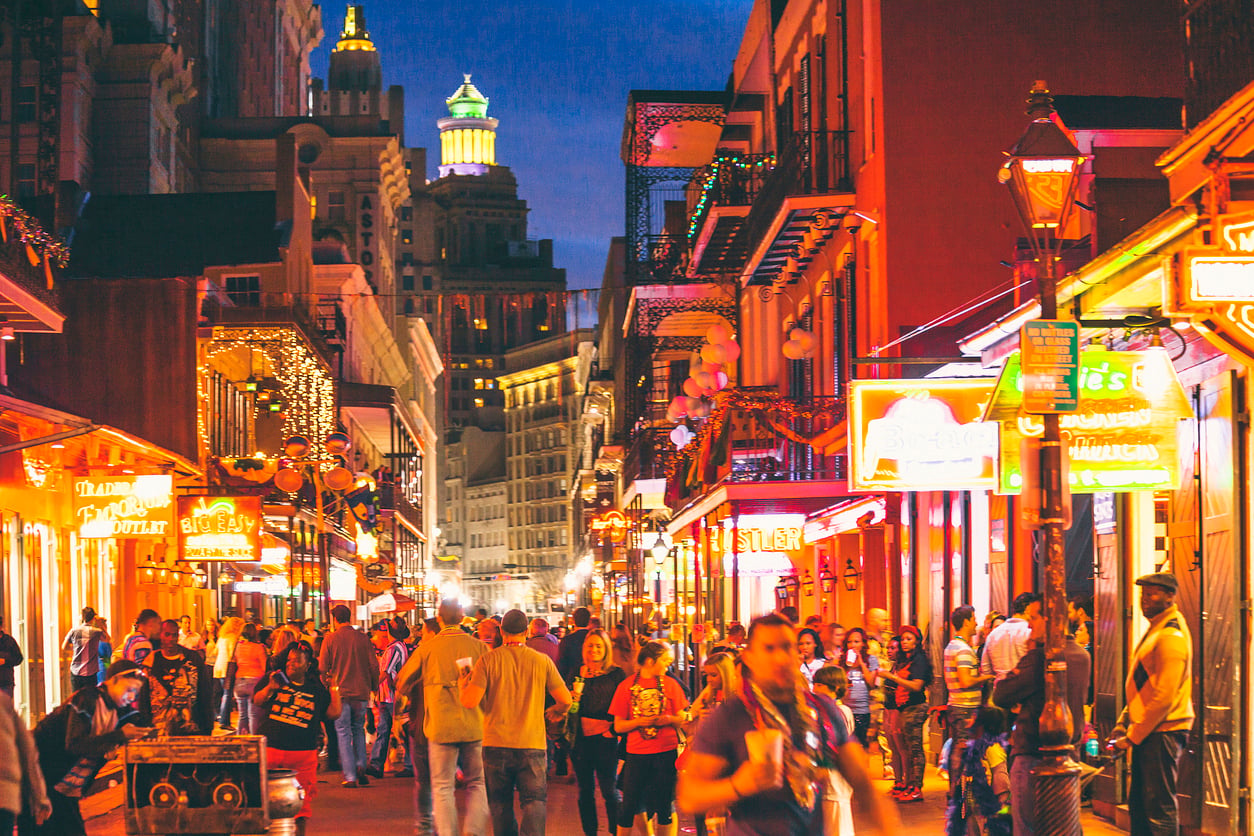 A vibrant, crowded street in New Orleans at night, filled with people, bright neon signs, and glowing lights from bars and restaurants, with tall buildings in the background.