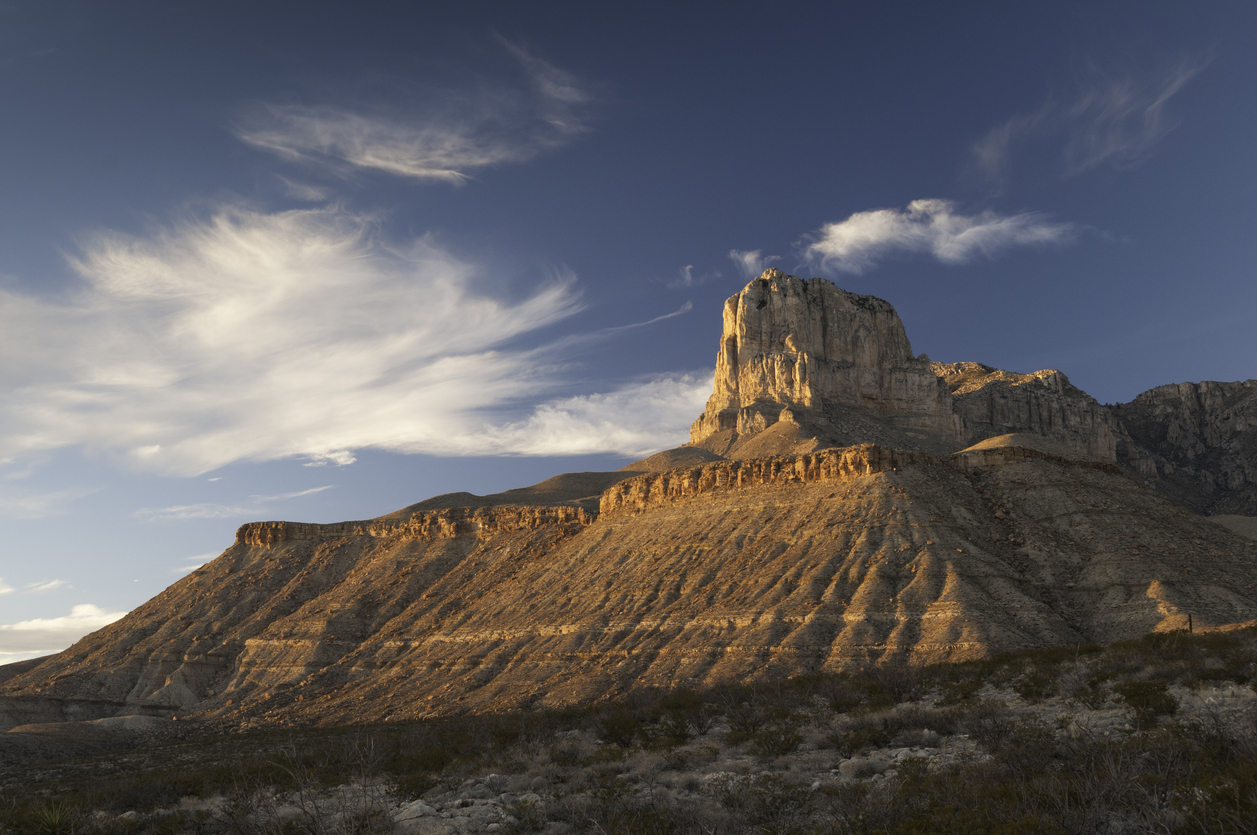 A rocky mountain peak rises above layered cliffs, bathed in warm sunlight with wispy clouds in a blue sky. Renowned as one of the top hiking destinations, the area’s desert vegetation covers the lower foreground.