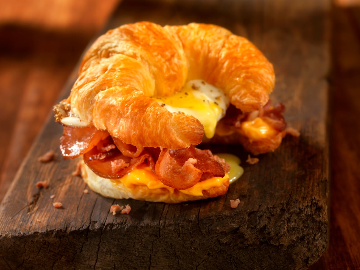 A croissant sandwich filled with crispy bacon, melted cheese, and a fried egg with runny yolk, served on a rustic wooden board.