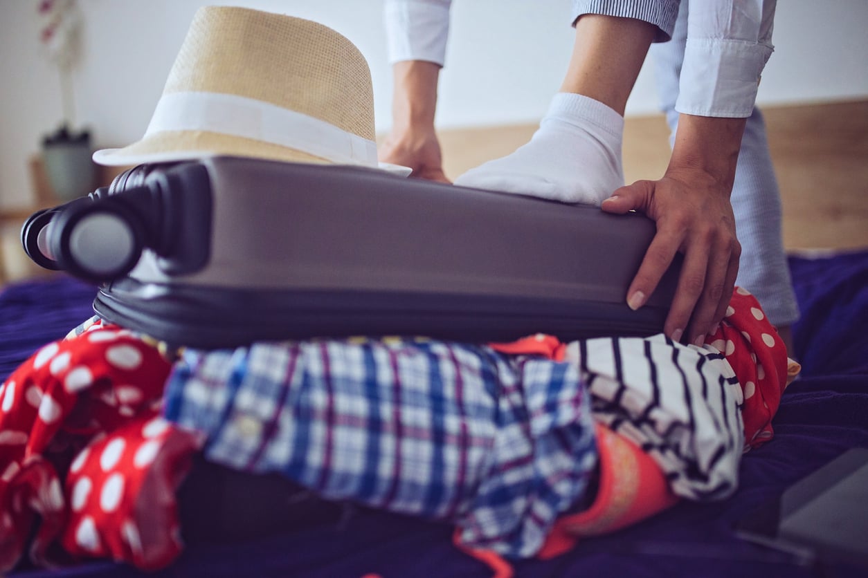 A person presses down on an overstuffed suitcase with one hand and a foot while trying to close it; clothes spill out, and a hat rests on top.