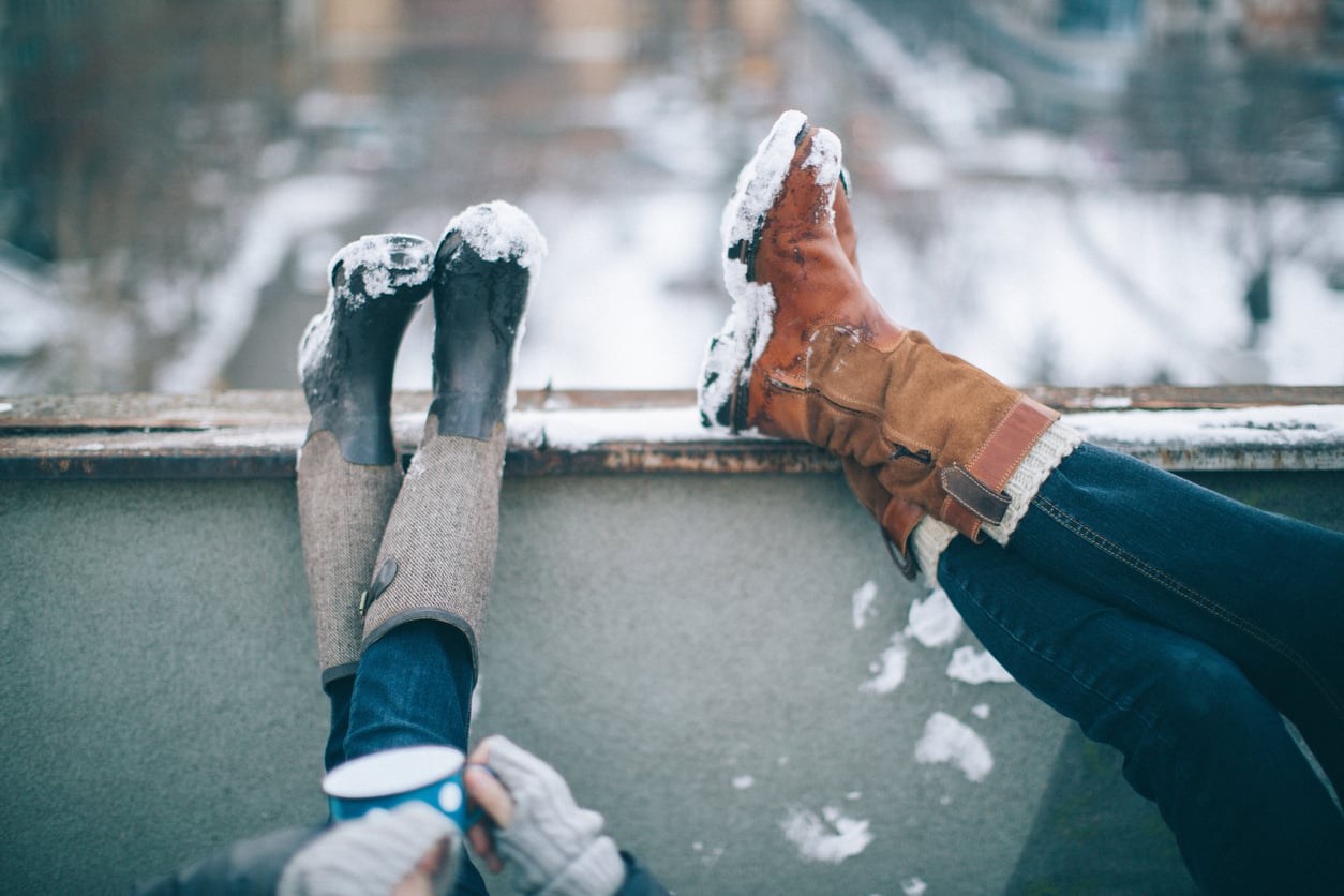 Two people sitting outdoors with snowy boots propped up on a ledge. One person wears brown boots; the other wears black. The scene is wintry with blurred snow-covered buildings and streets in the background.