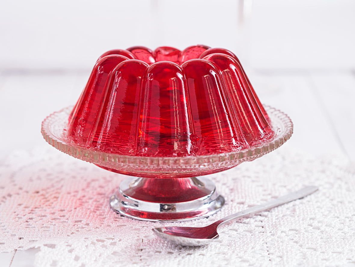 A red gelatin dessert sits on a glass pedestal dish, placed on a white lace tablecloth, with a silver spoon beside it.