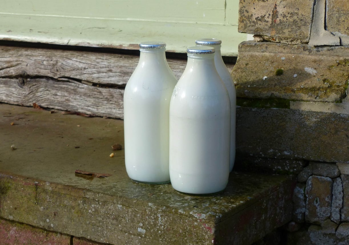 Four glass milk bottles with metal caps are placed on a stone doorstep next to a wall and wooden planks in an outdoor setting.