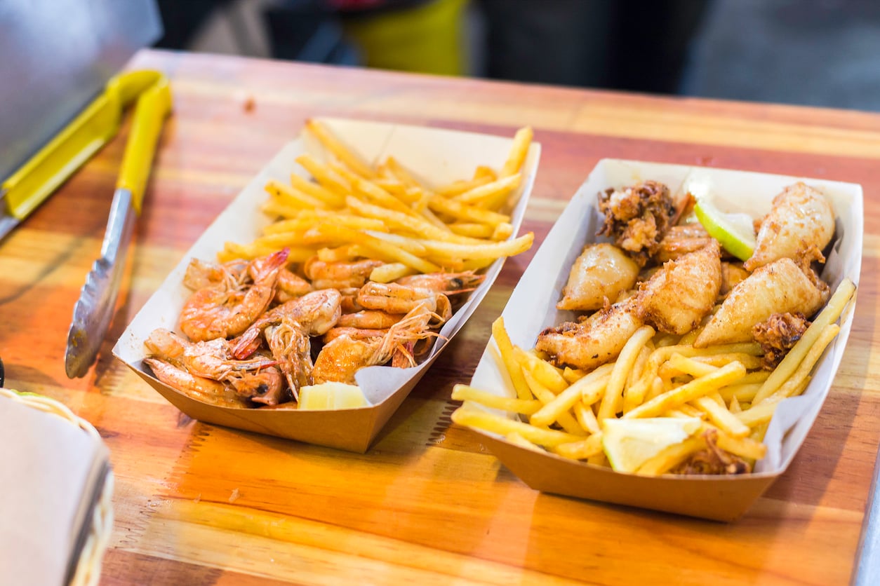 Two cardboard trays on a wooden table, one with fried shrimp and French fries, the other with fried fish pieces, lemon wedges, and fries. Metal tongs and a napkin are nearby.