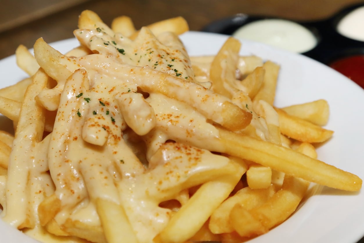 French fries topped with creamy cheese sauce and sprinkled with seasoning, served on a white plate. Dipping sauces are visible in the background.