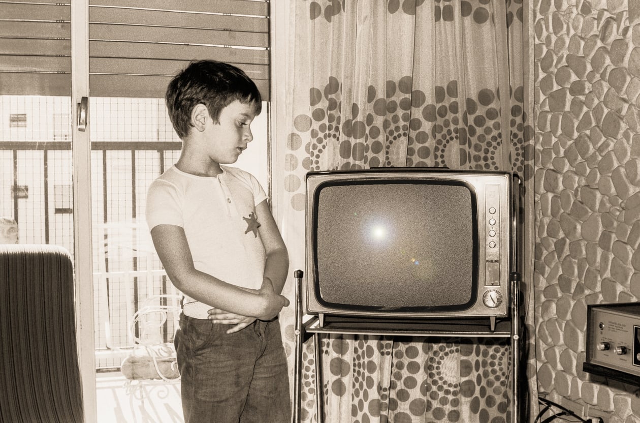 A young boy stands beside an old-fashioned television in a vintage living room, looking at the TV screen. A patterned curtain and window are in the background.