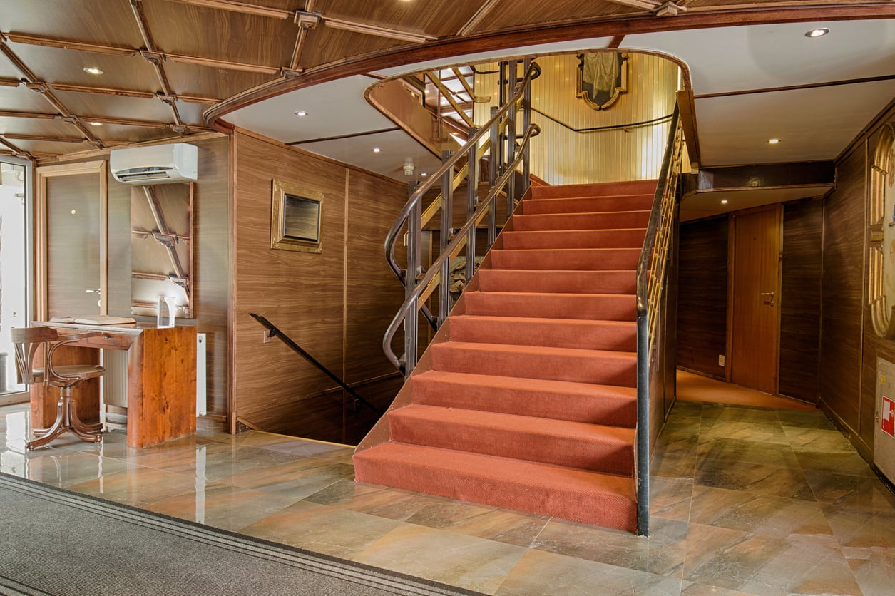 A modern interior with a red-carpeted staircase, wooden walls, tiled floor, and a small desk with two chairs. Warm lighting and a door are visible in the background.