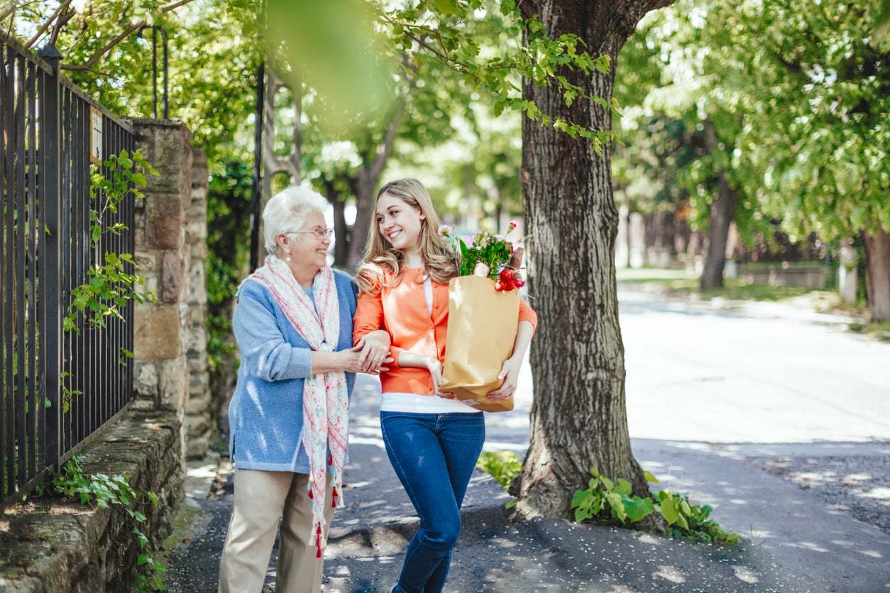 A young woman carrying groceries smiles while walking arm-in-arm with an elderly woman on a sunny, tree-lined sidewalk.