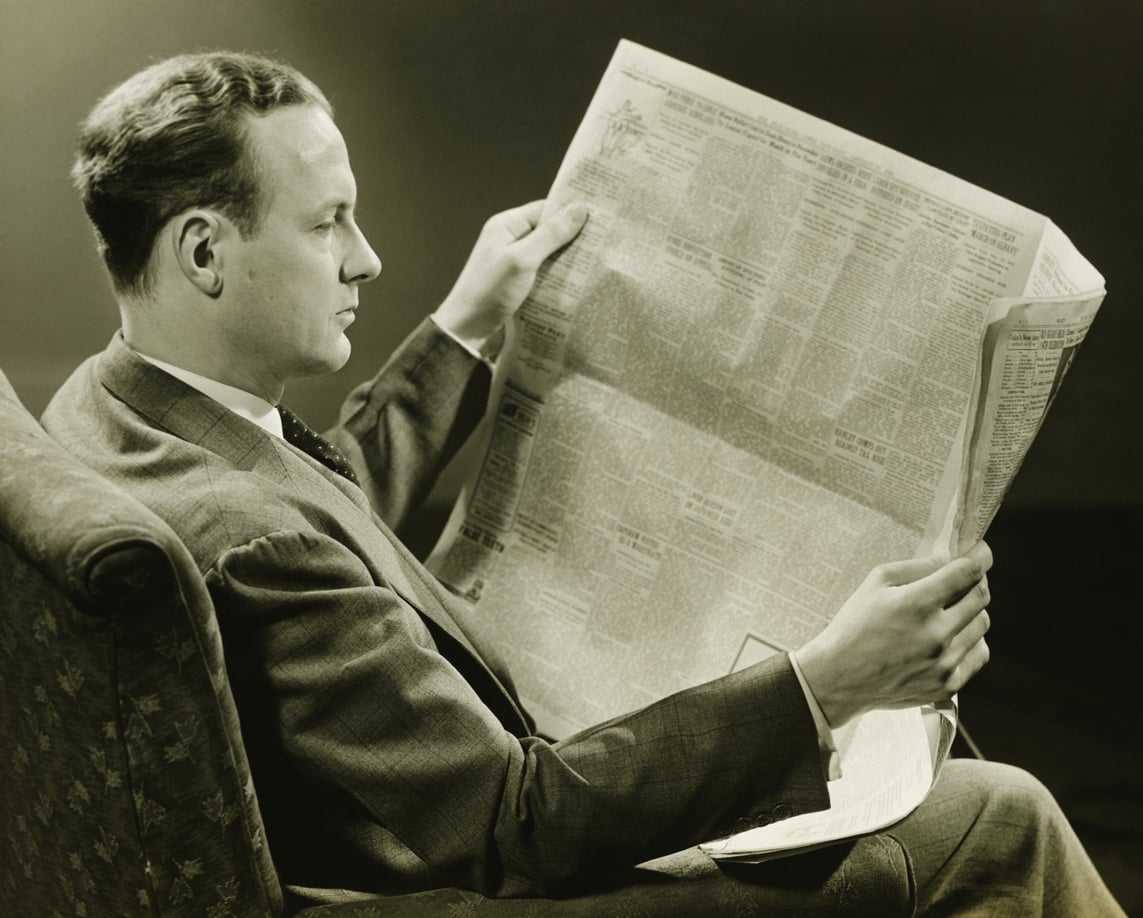 A man in a suit sits in a chair, reading a large newspaper. The image is black and white, and he appears focused on the newspaper’s content.