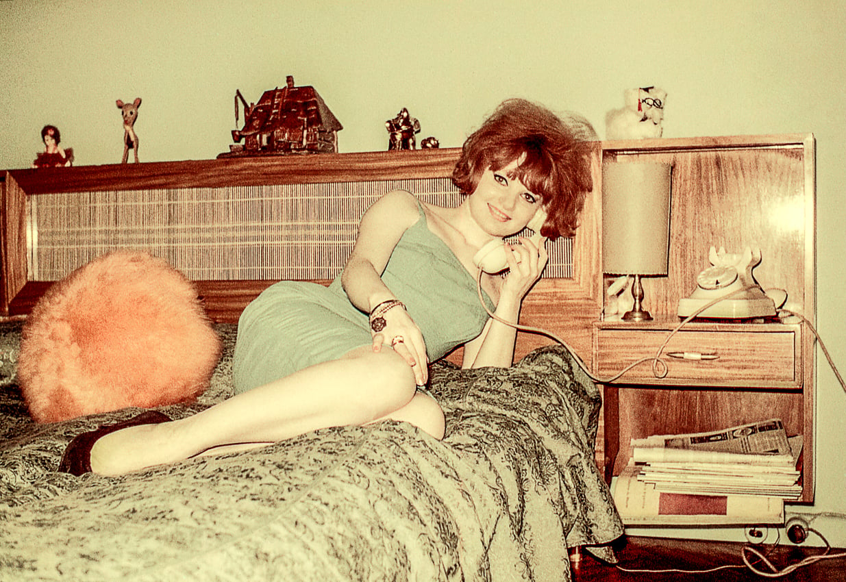 A woman in a green dress lounges on a vintage bed, talking on a rotary phone. The room features retro decor, a wooden headboard with figurines, a lamp, and a fluffy orange pillow.