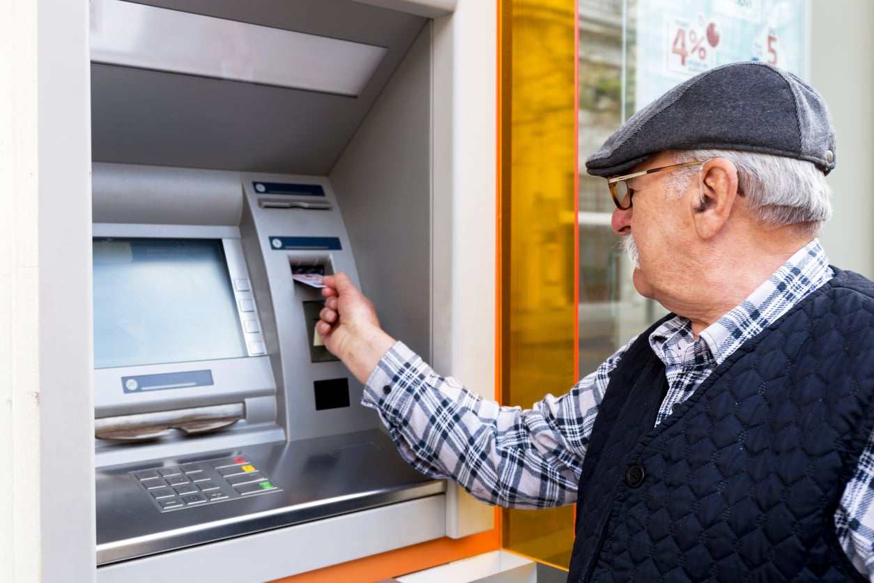 An elderly man wearing a flat cap and glasses uses an ATM, demonstrating prudent retirement money habits as he holds a bank card in his right hand while standing in front of the machine.