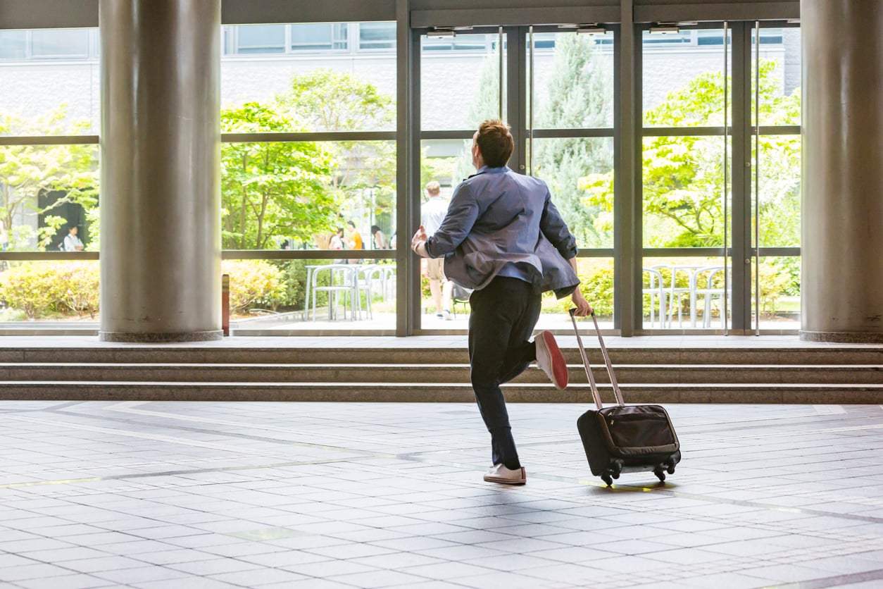 A man in a blue suit runs through an airport or transit terminal, pulling a small rolling suitcase behind him, heading toward large glass doors with greenery visible outside.